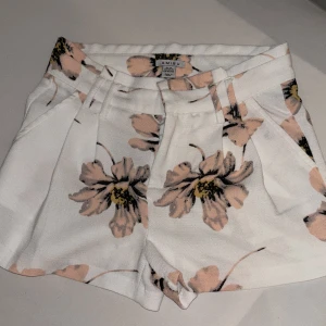 Blommiga vita shorts från Amisu - Snygga vita shorts från Amisu med stort blommigt mönster i ljusrosa och svart. Modellen har hög midja, bälteshällor och fickor både fram och bak. Materialet är mjukt och känns som bomull, perfekt för varma dagar. Passformen är normal och de har en fräsch, somrig vibe.