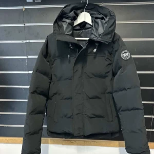 Svart pufferjacka med huva - Säljer en svart pufferjacka från Canada Goose med huva och patch på ärmen. Jackan har dragkedja och snörning vid halsen, samt två stora fickor framtill. Perfekt för kalla dagar och riktigt snygg stil.