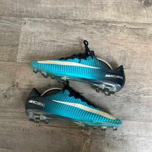 Nike Mercurial blå fotbollsskor 45 - Säljer ett par Nike Mercurial fotbollsskor i blått med svarta och vita detaljer. Skorna har snörning, platt sula med dobbar och ett randigt mönster. Swoosh-loggan syns tydligt på sidan och sulan har en metallic finish. Perfekt för dig som vill sticka ut på planen.