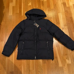 Polo Ralph Lauren puffer jacket - Snygg svart pufferjacka från Polo Ralph Lauren med huva och två sidofickor med dragkedja. Jackan har en röd broderad logga på bröstet och quiltad design. Denna jacka är perfekt nu till vinter när det är kallt ute.