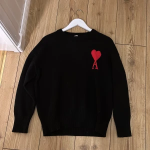 Svart stickad tröja från Ami Paris - Svart stickad tröja från Ami Paris med röd hjärtlogga och bokstaven A på bröstet. Tröjan har rund hals, ribbade muddar och lång ärm. Perfekt för dig som gillar stilrena plagg med en twist. Materialet är mjukt och håller värmen bra.