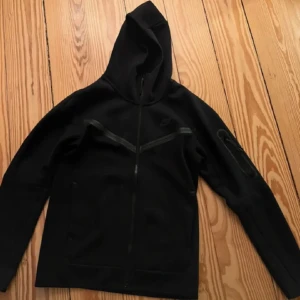 Svart Nike hoodie med dragkedja - Svart hoodie från Nike med hel dragkedja och huva. Tröjan har långa ärmar och snygga detaljer med svarta linjer över bröstet samt en ficka med dragkedja på ena ärmen. Perfekt för chill eller träning, stilren och sportig look.