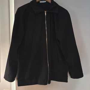 Svart jacka med dragkedja från Kumishi - Stilren svart jacka från Kumishi med rak passform och klassisk krage. Jackan har hel dragkedja framtill och två sidofickor. Materialet är bomull och jackan är perfekt för dig som gillar en clean look.