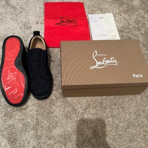 Svarta sneakers från Christian Louboutin - Snygga svarta sneakers från Christian Louboutin i mocka med klassisk röd sula och rund tå. Skorna har svarta snören och diskreta metalldetaljer på sidan. Perfekta för dig som vill ha en lyxig och stilren look med en ikonisk touch. Använda en gång kan bytas 
