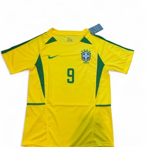 Brasilien Ronaldo 9 fotbollströja Nike - Gul Brasilien fotbollströja med gröna detaljer, nummer 9 och Ronaldo på ryggen. Nike-logga och landslagsemblem på bröstet. Tillverkad i lätt och ventilerande polyester med Dri-Fit teknologi för att hålla dig torr. Kortärmad och klassisk passform.