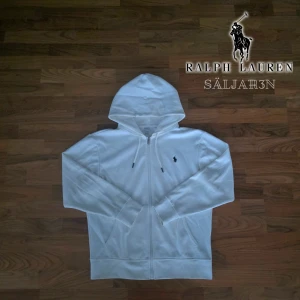 Vit Ralph Lauren Hoodie - Vit hoodie från Polo Ralph Lauren med dragkedja och huva. Klassisk design med svart broderad logga på bröstet, snörning i huvan och två fickor framtill. Tillverkad i mjuk bomull som är skön att ha på sig. Perfekt för chill dagar och streetwear.