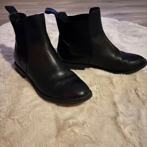 Svarta chelsea boots i skinn stl 37 - Snygga svarta chelsea boots med rund tå och platt sula. Bootsens ovandel är i slätt skinn och har elastiska paneler på sidorna för enkel på- och avtagning. Klassisk siluett med dragflik bak och låg klack, perfekta för en stilren look.