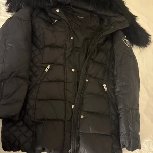 Svart pufferjacka med fuskpäls - Säljer en svart pufferjacka med quiltade detaljer och fuskpäls på huvan. Jackan har dragkedjor, knappar och ett snyggt emblem på ärmen. Perfekt för kalla dagar och har en modern, lite oversized passform.