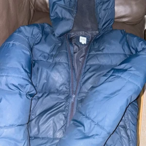 Blå pufferjacka från Hugo Boss - Säljer en blå pufferjacka från Hugo Boss med huva och dragkedja. Jackan har quiltad design och är tillverkad i syntetmaterial, perfekt för kalla dagar. Stilsäker look med diskret logga på insidan och klassisk passform.