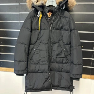 Svart Parajumpers dunjacka med päls - Svart lång dunjacka från Parajumpers med fluffig päls på huvan, cool gul detalj vid halsen och patch på ärmen. Jackan har dragkedja, knappar och två stora fickor framtill. Perfekt för kalla dagar och har en riktigt snygg streetstil.
