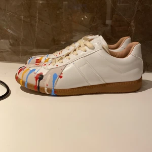 Maison Margiela sneakers med färgstänk - Unika vita sneakers från Maison Margiela med coola färgstänk i blått, rött, gult och svart på tåpartiet. Skorna har snörning, rund tå och en brun gummisula. Tillverkade i skinn och mocka för en lyxig känsla och edgy look.