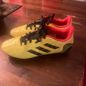 Adidas Copa gula fotbollsskor - Säljer ett par Adidas Copa fotbollsskor i en cool gul färg med svarta detaljer och röd insida. Skorna har klassisk snörning och mönstrad ovandel i syntetmaterial. Yttersulan har fasta dobbar för gräsplan. Perfekt för dig som vill sticka ut på planen.