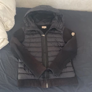 Svart pufferjacka från Moncler S - Svart pufferjacka från Moncler med stickade ärmar och huva. Jackan har quiltad kropp, dragkedja framtill och en ficka med dragkedja på ärmen. Moncler-logga på vänster ärm. Snygg, modern look med blandat material för extra stilpoäng.