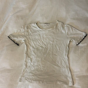 Vit t-shirt med spetsdetalj - vit t-shirt med korta ärmar och svart spetsdetalj vid ärmsluten. T-shirten har en klassisk rund hals och är gjord i mjukt bomullsmaterial