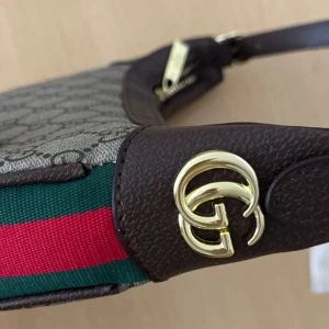 Gucci axelväska med GG-mönster - Snygg axelväska från Gucci med klassiskt GG-mönster i beige och brunt. Väskan har bruna skinn-detaljer, guldfärgad GG-logga och dragkedja. Grön och röd rand på sidan ger den ikoniska Gucci-looken. Perfekt storlek för det viktigaste.