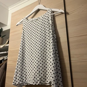 Vit prickig linnetopp med knapp - Säljer en vit linnetopp med svarta prickar och rund hals. Toppen är ärmlös och har en liten knapp i nacken för enkel påklädning. Materialet känns mjukt och lätt. Passformen är normal och toppen har en rak, luftig siluett.