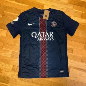 PSG fotbollströja Nike blå röd - Snygg PSG fotbollströja från Nike i mörkblått med röd och vit randig detalj på framsidan. Tröjan har korta ärmar, klubbmärke på bröstet och Qatar Airways tryck. Tillverkad i lätt och ventilerande material, perfekt för match eller träning.