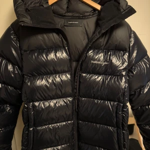 Svart Peak Performance pufferjacka XL - Säljer en svart pufferjacka från Peak Performance i storlek XL. Jackan har en glansig finish, huva och tydliga quiltade sektioner. Den stängs med dragkedja och har en stilren look med diskret logga på bröstet. Perfekt för kalla dagar.