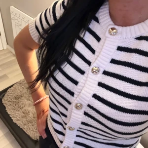 Randig vit och svart kofta med kort ärm - Säljer en snygg kortärmad kofta i vit och svart med randigt mönster. Tröjan har silvriga knappar framtill och är stickad med ribbad kant vid hals och knappslå. Perfekt för dig som gillar en fräsch och trendig look.