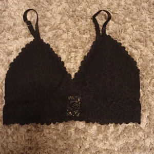 Svart spetsig bralette från Lager 157 - Svart bralette från Lager 157 i storlek XS, med spetsdetaljer och justerbara axelband. Toppen har en snygg blommig spets och är ärmlös med en triangelformad kupa. Aldrig använd.