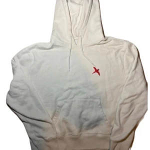 Biege hoodie med röd fågelbrodyr - Vit hoodie med snörning i huvan och en broderad röd fågel på bröstet. Tröjan har en klassisk känguruficka framtill och ribbade muddar vid ärmslut och nederkant. Perfekt för dig som gillar stilrena detaljer och vill ha en chill look.