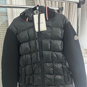Svart Moncler pufferjacka med huva - Svart pufferjacka från Moncler med quiltad front och stickade ärmar. Jackan har huva med snörning och coola rödvita detaljer på kragen. Moncler-logga på ärmen och dragkedja framtill. Perfekt för dig som vill ha en snygg och varm jacka med street vibe.