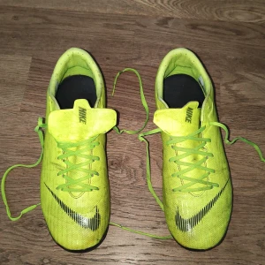 Nike Mercurial fotbollsskor gul - Nike Mercurial fotbollsskor i en skarp gul färg med svarta detaljer och logga. Skorna har snörning, rund tå och platt sula med dobbar för gräsplan. Tillverkade i syntetmaterial för lätt känsla och bra grepp. Perfekt för dig som vill sticka ut på planen.