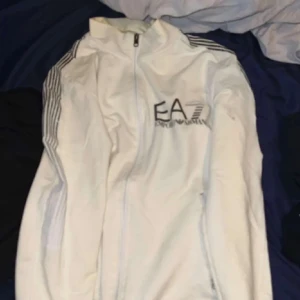 Vit EA7 vindjacka med dragkedja - Säljer en fräsch vit vindjacka från EA7 Emporio Armani med svarta ränder på axlarna och logga på bröstet. Jackan har hel dragkedja framtill. 