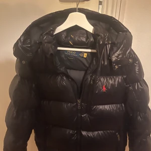 Svart dunjacka från Polo Ralph Lauren - Säljer en svart, glansig dunjacka från Polo Ralph Lauren med huva och dragkedja framtill. Köpt förra året på ralphlauren för 6000kr kvitto finns. Jackan har två små hål på innersidan av ärmarna inget som syns eller stör.