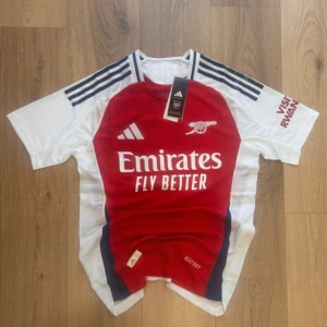 Arsenal Saka 7 matchtröja Adidas M - Arsenal hemmatröja med Saka 7 på ryggen, officiell Adidas-produkt i slim fit. Röd front med vita ärmar och mörkblå detaljer, klassiska Adidas-ränder på axlarna. Materialet är lätt och ventilerande polyester, perfekt för match eller träning. Emirates och Visit Rwanda-loggor tryckta.