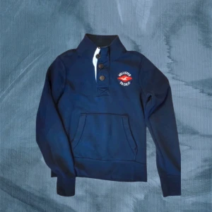 Marinblå Hollister kofta - Snygg marinblå hoodie från Hollister med hög krage och tre knappar framtill. Tröjan har en stor magficka och broderad logga på bröstet. Perfekt för dig som gillar en sportig och avslappnad stil.