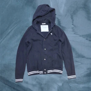 Mörkblå kofta Abercrombie & Fitch - Snygg mörkblå vindjacka från Abercrombie & Fitch med huva och vita ränder på muddar och nederkant. Jackan har knappar framtill, två fickor och en broderad vit älg på bröstet. Perfekt för dig som gillar sportig och clean stil.