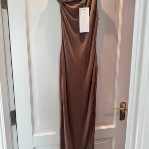 Brun långklänning från Johansen - Supersnygg brun långklänning från Johansen med twistade axelband och draperad överdel. Klänningen har en elegant, figurnära passform och är gjord i ett mjukt, skimrande material som ger en lyxig känsla. Perfekt för dig som vill sticka ut med stil.