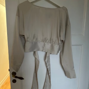 Beige omlottopp från Vero Moda - Säljer en beige omlottopp från Vero Moda med långa ärmar och breda manschetter med knappar. Toppen har ett snyggt omlottband i midjan som kan knytas och en öppen rygg för extra stilpoäng. Materialet är mjukt och har en lätt glansig finish.