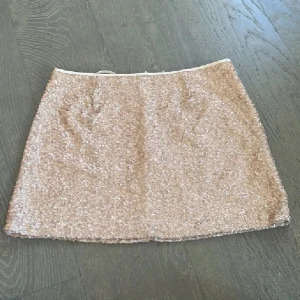 Champagnefärgad paljettkjol Jeane Blush - Snygg kortkjol från Jeane Blush i champagnefärgad ton, täckt med glittriga paljetter. Kjolen har en rak passform och dragkedja bak för enkel påklädning. Perfekt för dig som vill sticka ut med lite extra glitter.