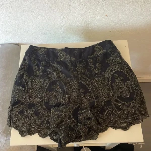 Svarta shorts med guldpaljetter - Unika svarta shorts med glittriga guldpaljetter och broderade detaljer. De har hög midja och ett snyggt scallop-mönster längs bensluten. Materialet är mjukt med ett innerfoder för extra komfort. Perfekt för dig som vill sticka ut med en glamorös vibe.