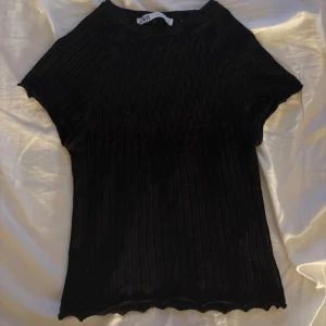 Svart ribbad topp från H&M - Svart ribbad topp från H&M med korta ärmar och rund halsringning. Toppens material är stretchigt och har en lätt transparent känsla. Nederkanten har en vågig finish som ger en extra detalj till looken.