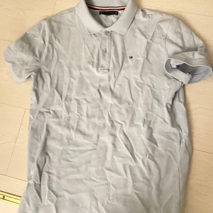 Ljusblå pikétröja Tommy Hilfiger - Snygg ljusgrå pikétröja från Tommy Hilfiger med klassisk krage och knäppning framtill. Tröjan har korta ärmar och den ikoniska lilla loggan broderad på bröstet. Perfekt för en clean och stilren look.