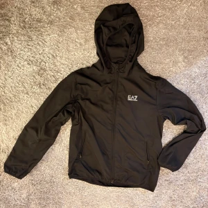 Svart EA7 vindjacka med huva - Svart vindjacka från EA7 Emporio Armani med stilren design och huva. Jackan har dragkedja framtill, två sidofickor med dragkedja och meshfoder på insidan. EA7-logga på bröstet ger en sportig vibe. Perfekt för dig som gillar clean och enkel stil. Jag har köpt jackan för en månad och jag använde den 2-3 gånger bara så den är typ helt ny. Köpte den för 1900kr och vill sälja den för mindre.