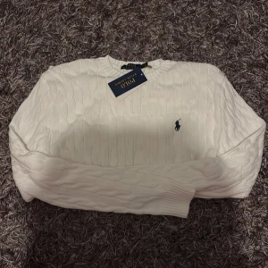 Vit kabelstickad tröja från Polo Ralph Lauren - Vit kabelstickad tröja från Polo Ralph Lauren med klassisk rund halsringning och broderad marinblå logga på bröstet. Tröjan har långa ärmar och ribbade muddar för en snygg passform. Perfekt för dig som gillar stilrena och tidlösa plagg.