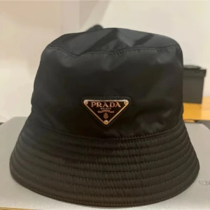 Svart bucket hat från Prada - Snygg svart bucket hat från Prada med klassisk triangulär logga framtill. Hatten har en stilren design i glansigt syntetmaterial och bred, nedåtlutande brätte. Perfekt accessoar för en trendig streetstyle-look.