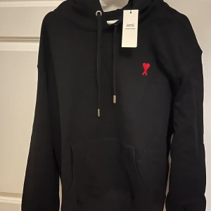 Svart hoodie från Ami med rött hjärta - Svart hoodie från Ami Alexandre Mattiussi med klassisk huva, snörning och stor känguruficka. På bröstet finns Ami:s ikoniska röda hjärta med A-broderi. Perfekt för en stilren och avslappnad look. Passar storlek M upp till L