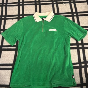 Grön pikétröja i frotté från Stüssy - Säljer en grön pikétröja från Stüssy i mjuk frotté. Tröjan har vit krage med svarta ränder, half zip-dragkedja och en liten ficka med vit kant på bröstet. Perfekt för dig som gillar retrovibbar och streetwear.