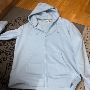 Ljusblå hoodie från Lacoste - Snygg ljusblå hoodie från Lacoste med dragkedja och klassisk krokodillogga på bröstet. Tröjan har huva med snörning, två stora fickor framtill och är i mjuk bomull. Perfekt för chill dagar eller när du vill ha en clean look.