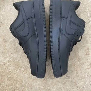 Nike Air Force 1 helsvarta sneakers - Säljer ett par klassiska Nike Air Force 1 i helsvart skinn. Skorna har låg profil, rund tå, snörning och den ikoniska Air-sulan. Perfekta för dig som gillar en clean och stilren look med diskreta detaljer och AF1-märke på snörningen.