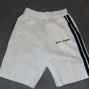 Vita Palm Angels shorts med svarta ränder - Palm Angels vita shorts med svarta sidoränder och broderad logga på vänster ben. De har elastisk midja och raka ben för en clean och sportig vibe. Perfekta för dig som gillar streetwear och vill sticka ut med en klassisk look.