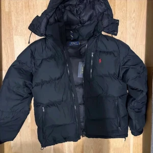 Svart dunjacka från Polo Ralph Lauren - Svart dunjacka från Polo Ralph Lauren med röd logga på bröstet. Jackan har huva, flera dragkedjeförsedda fickor och quiltad design. Perfekt för kalla dagar med sin tjocka och puffiga look. Snygg och stilren, passar dig som gillar klassisk streetwear.