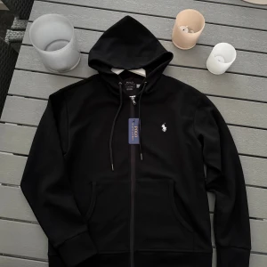 Svart hoodie från Polo Ralph Lauren - Svart hoodie från Polo Ralph Lauren med klassisk vit broderad logga på bröstet. Tröjan har huva med snörning, hel dragkedja framtill och två fickor. Materialet är mjuk bomull och passformen är normal. Perfekt för chill dagar och streetwear.