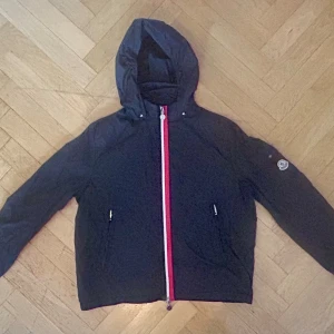 Svart vindjacka från Moncler med huva - Svart vindjacka från Moncler med huva och snygg dragkedja i rött, vitt och blått. Jackan har två fickor med dragkedja och ficka med Moncler-logga på ärmen. Luvan går att veckla in och stängas med tryckknappar. Perfekt för dig som gillar stilrena och sportiga plagg med exklusiv känsla.
