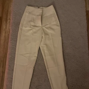 Beige kostymbyxor med hög midja - Snygga beige kostymbyxor med hög midja och raka ben. Byxorna är från Zara och aldrig använda. Har bara legat i garderoben, dom e super fina till både skor och klackar. Storlek Xs